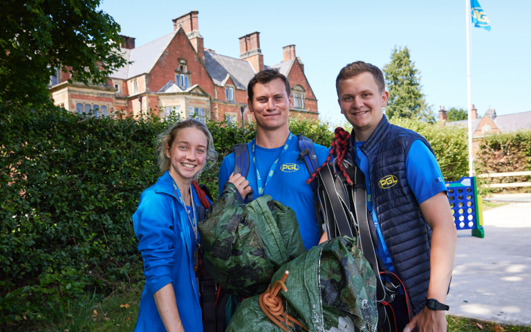 Three individuals in matching blue uniforms stand outside with equipment, smiling at the camera. A brick building and trees are visible in the background.