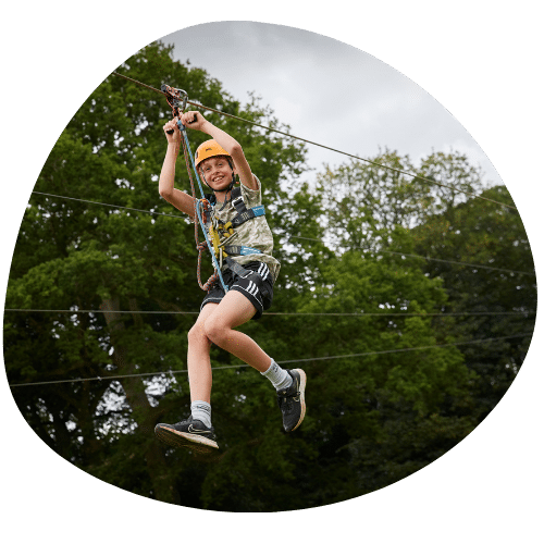 A person wearing a safety harness and helmet is riding along a zip line against a backdrop of trees.