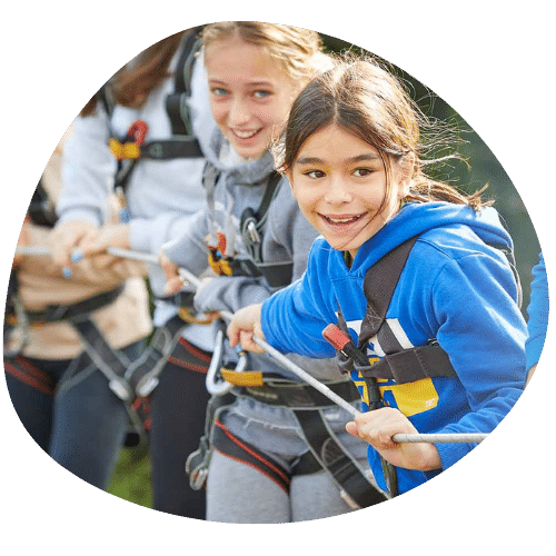 Children participate in an outdoor rope activity, wearing safety harnesses and smiling while pulling on a rope together.