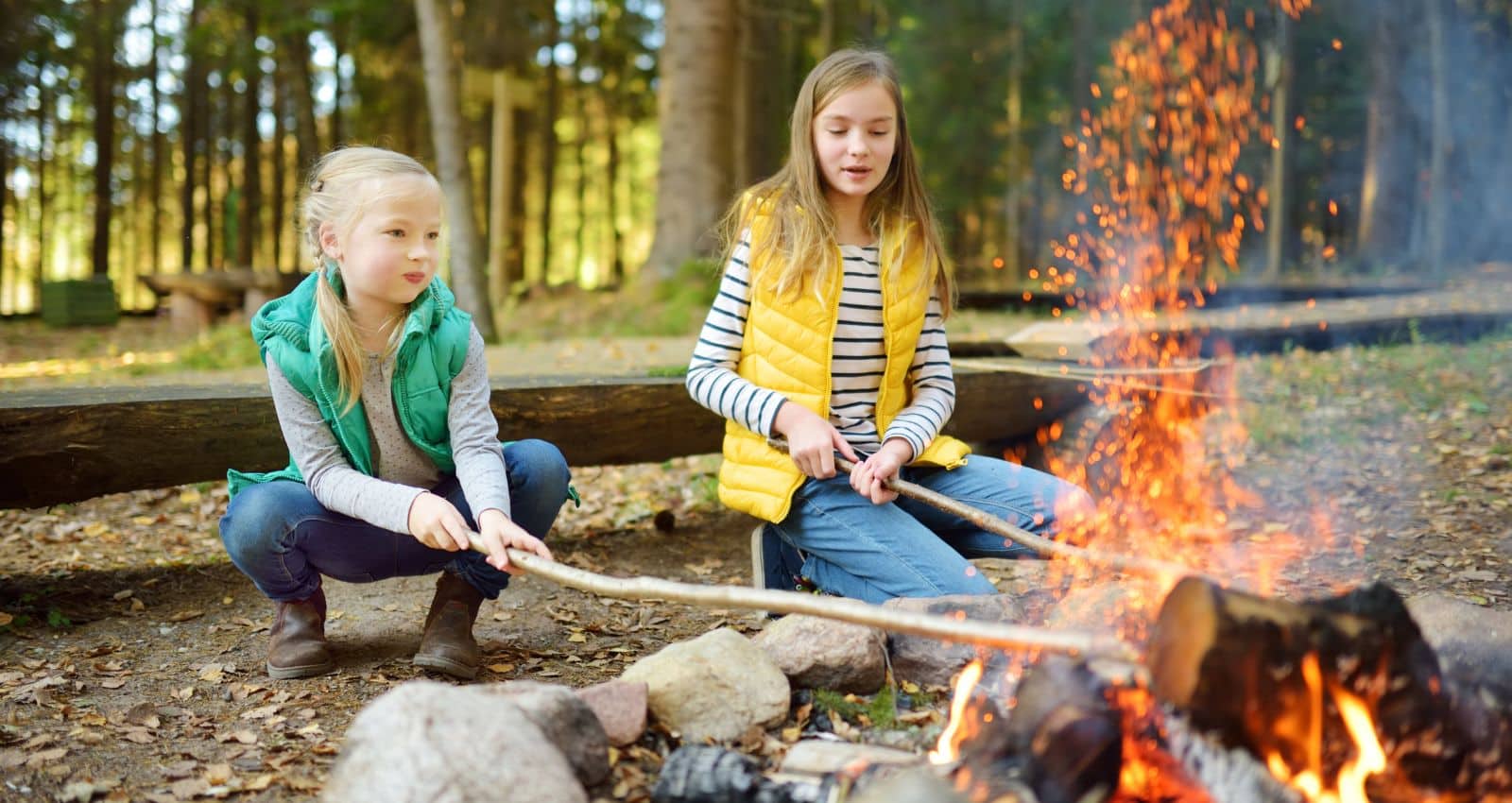 Two girls sitting by a campfire, roasting food on sticks in a forest setting. Both are wearing casual outdoor clothes, one in a green vest and the other in a yellow vest.