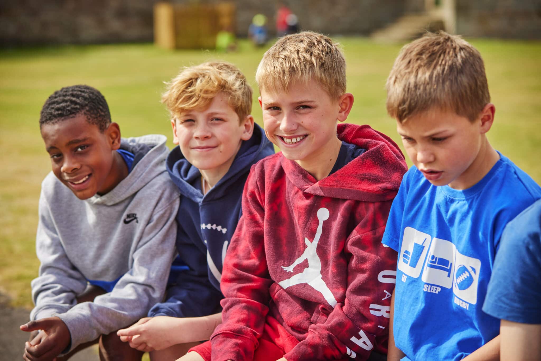 Four boys sit on a bench outside on a grassy field, smiling and looking at the camera. They wear casual clothes, including hoodies and t-shirts.