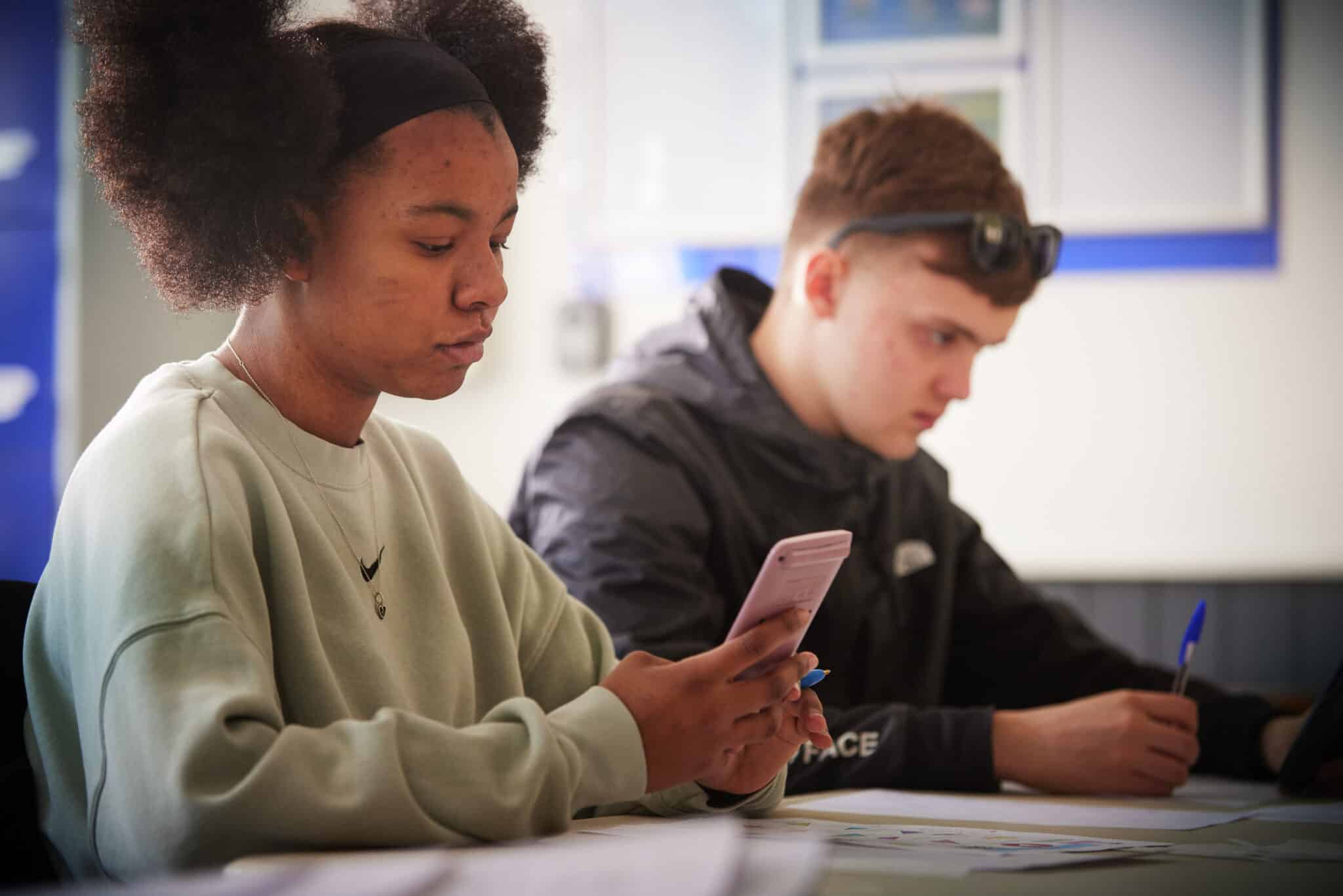 Two students are seated at a table, focused on their tasks. The student in the foreground is looking at a smartphone, while the student in the background is writing with a pen.