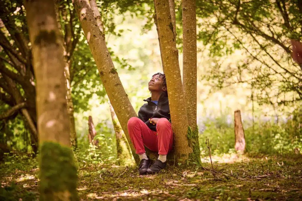 A person with short hair sits at the base of a tree in a forested area, looking up. They are wearing a dark jacket, red pants, and black shoes. Bright greenery surrounds the scene, suggesting they might be enjoying some multi-activity outdoor time.