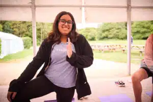 A person with glasses and a blue shirt is smiling and posing in a standing yoga position outdoors, under a canopy. The scene hints at a multi-activity space, with a picnic area and greenery in the background.