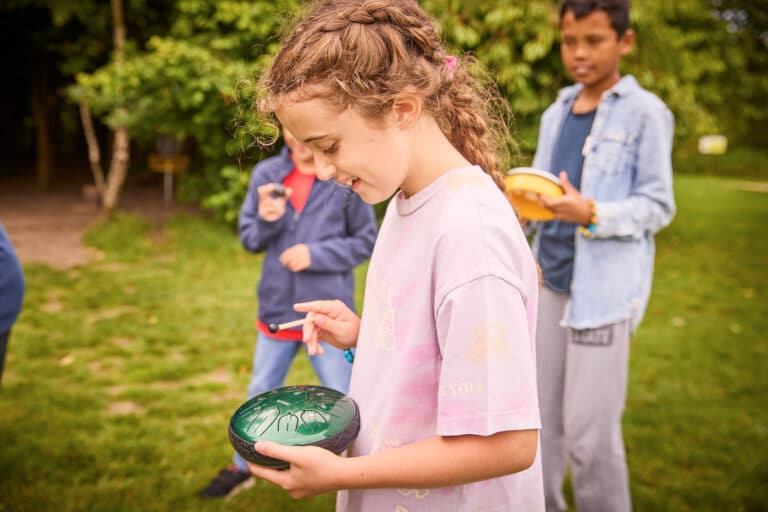 A young girl is holding a green object outdoors, surrounded by other children. They appear to be engaged in a multi-activity session in a grassy area with trees in the background.