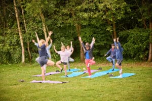 Children engaging in a multi-activity session are practicing yoga on mats in a grassy outdoor area with trees in the background. They are standing on one leg with arms raised, mimicking the tree pose.