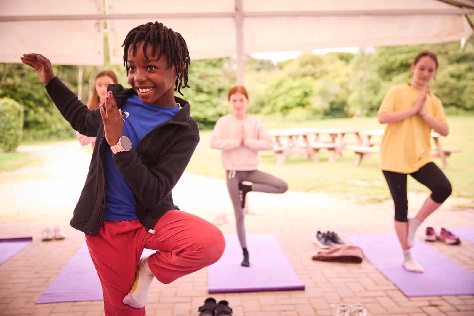 A group of four people practices yoga poses on outdoor mats, with the person in the front posing energetically and the three others behind holding tree poses. The background highlights a multi-activity picnic area surrounded by greenery.