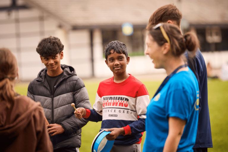 A group of people participates in a multi-activity event outdoors. Two boys hold instruments, and a woman in a blue shirt stands in the foreground. A half-timbered building is visible in the background.