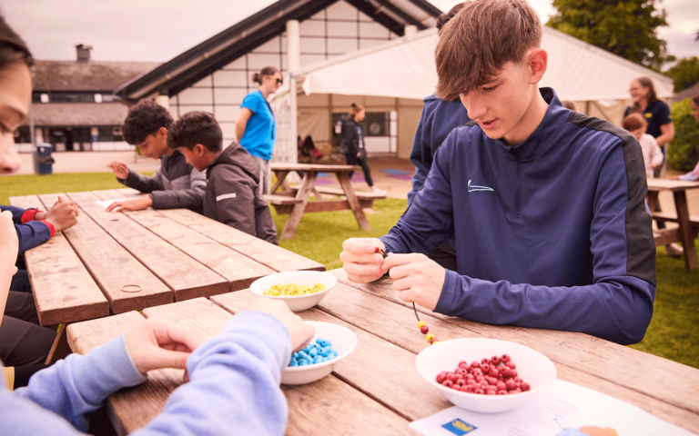 Teenagers seated outdoors at a wooden table, each working on a bead craft project, with bowls of colored beads and string materials in front of them. A building and a tent, suggesting a multi-activity space, are visible in the background.