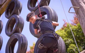 A person in a harness climbs a tire obstacle structure outdoors on a sunny day, surrounded by trees in the background and taking part in this multi-activity adventure.