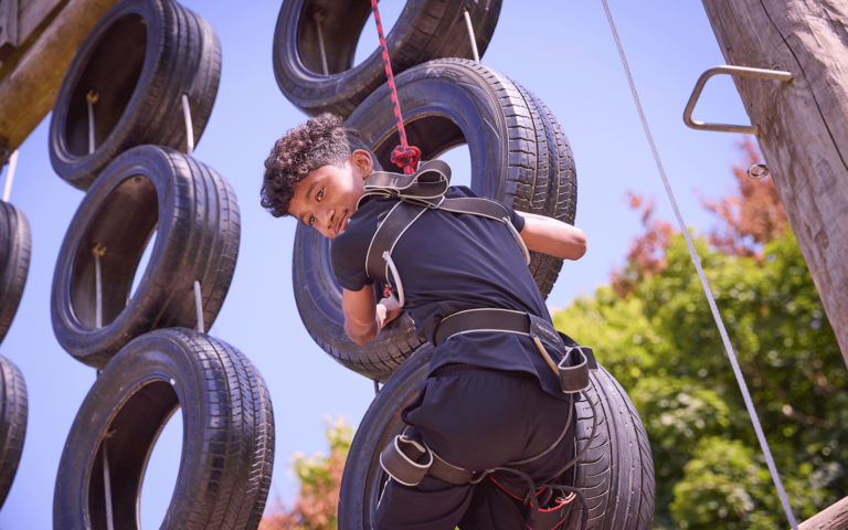 A person in a harness climbs a tire obstacle structure outdoors on a sunny day, surrounded by trees in the background and taking part in this multi-activity adventure.