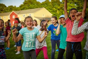 A group of children are playing energetically outdoors on grass, with colorful houses in the background. They are smiling and appear joyful, reminding one of the excitement found on outdoor school trips.