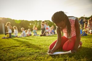 A child kneels on the grass, writing on paper, during an outdoor school trip, with a group of seated children in the background under a sunny sky.