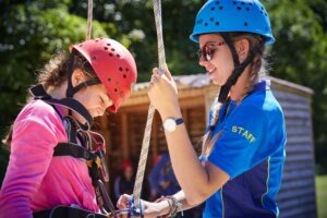 Two people wearing helmets, one in a safety harness, are outdoors. The person in a blue shirt assists the other with rope equipment.