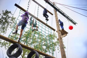 Three people wearing helmets navigate an outdoor ropes course featuring a tire obstacle, netting, and a red ball amidst the trees, offering a thrilling example of how to promote physical activity in schools.