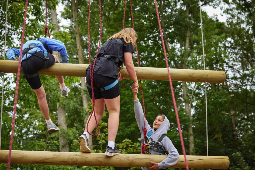 Three people participate in an outdoor ropes course, exemplifying how to promote physical activity in schools. Two are climbing large horizontal logs, while the third, in a hoodie, offers assistance.