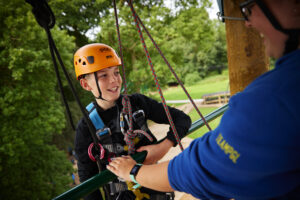 A person wearing a helmet and harness is secured with ropes by a staff member at this adventure facility, illustrating how promoting physical activity in schools can inspire an active lifestyle. Towering trees in the background complete the thrilling scene.