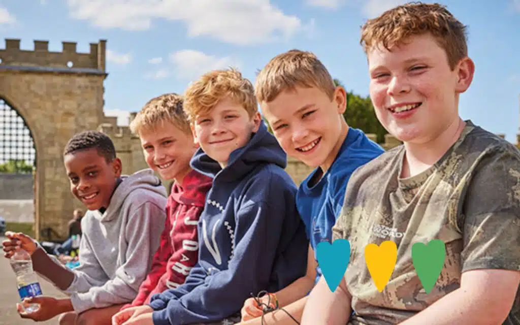 Five boys sitting on a ledge outdoors, wearing casual clothes and smiling. Stone archway and blue sky in the background.