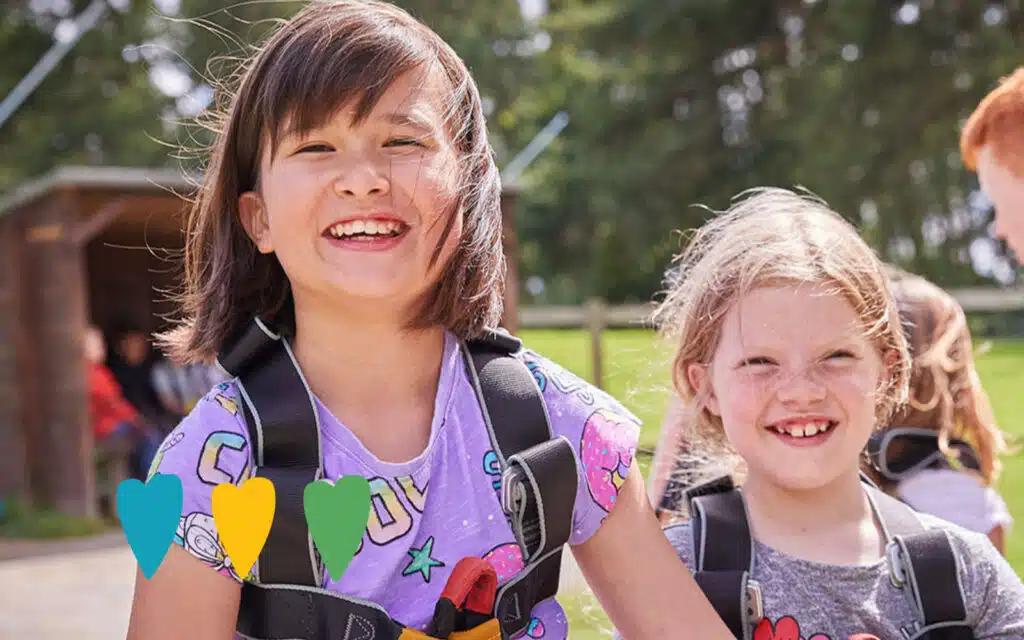 Two children wearing climbing harnesses smile outdoors, with greenery and a wooden structure in the background.