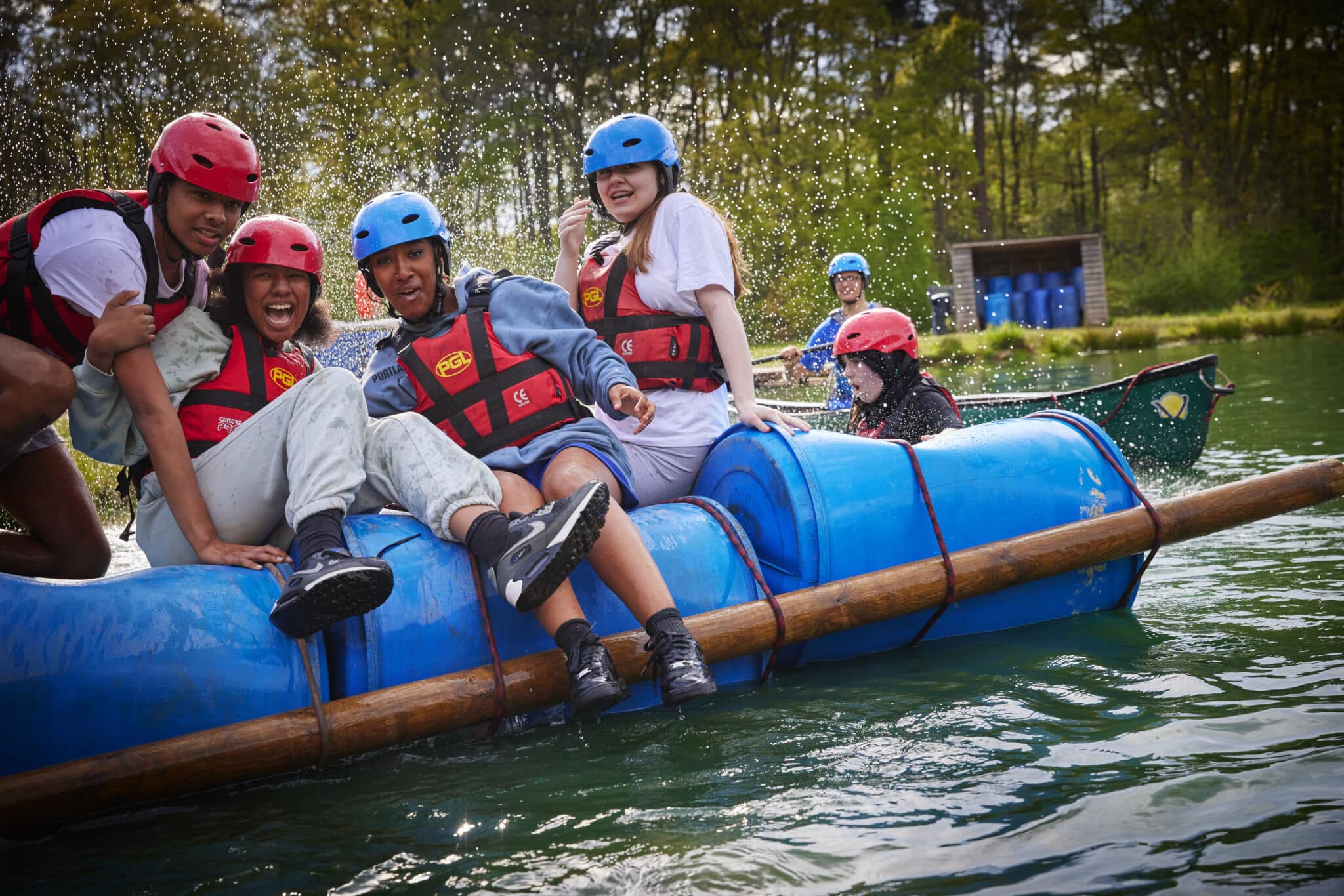 A group of people wearing helmets and life jackets are sitting on a blue raft made of barrels and logs, floating on a lake. Some water splashes around them. Trees and a small building are in the background.