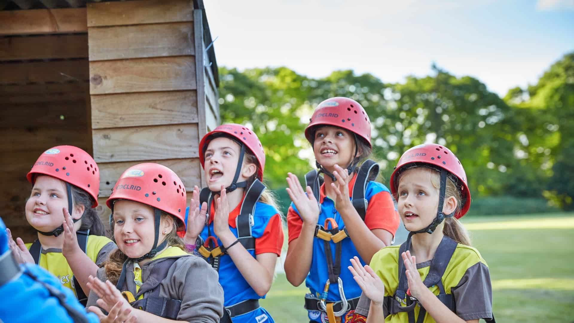 Children wearing helmets and harnesses stand outside, clapping and looking up. They are next to a wooden structure with trees and grass in the background.