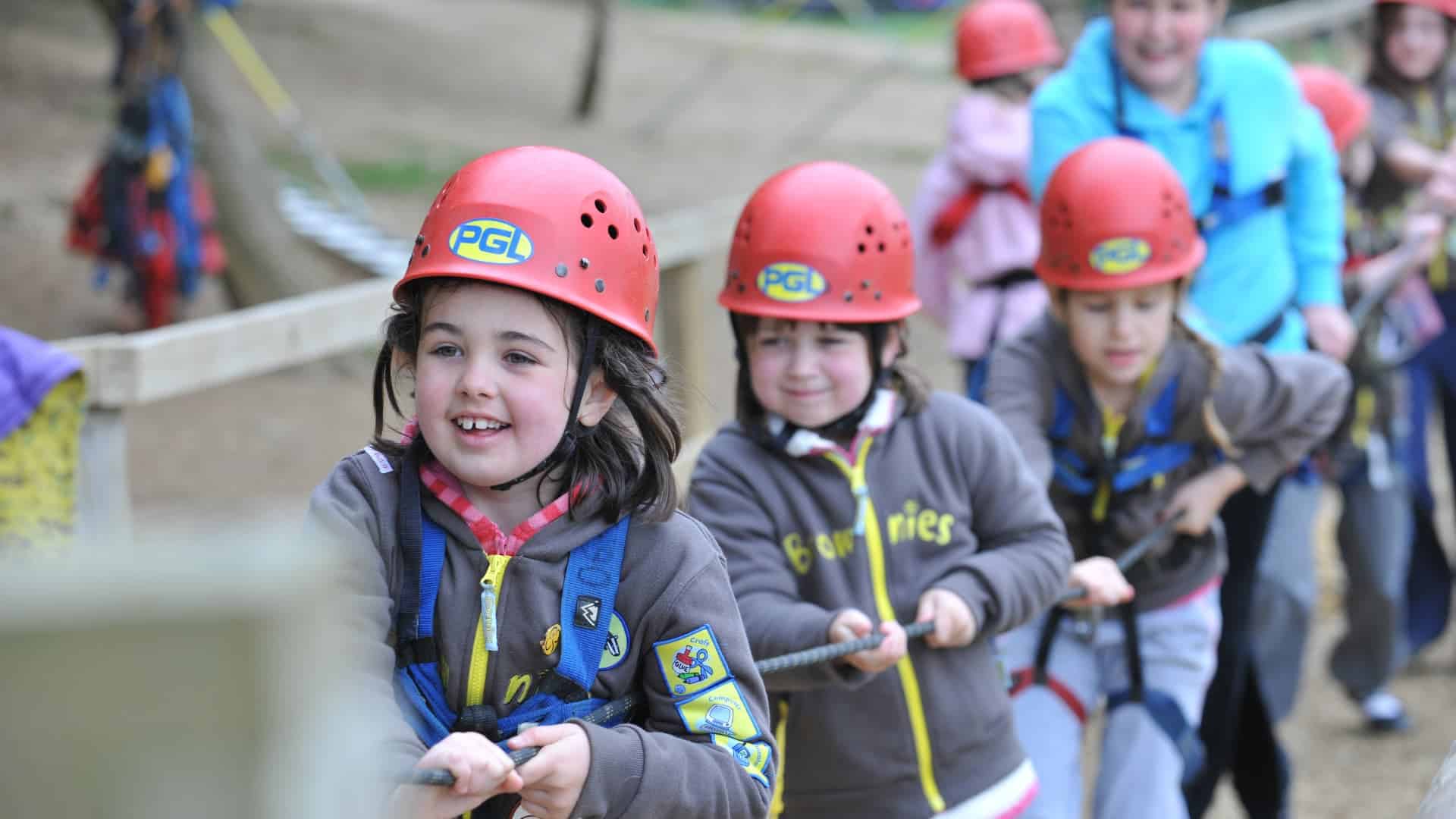 Children wearing helmets and harnesses participate in a teamwork activity involving pulling on a rope.