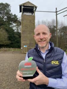 A person in a PGL vest holds an award outside, standing in front of a tall wooden structure.