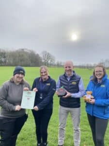 Four people stand in a grassy field holding a certificate, an award, and a stuffed bear. The sky is overcast.