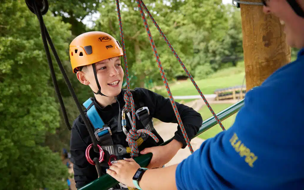 A boy in an orange helmet and harness stands on a platform, holding ropes, with a person in a blue shirt assisting him in an outdoor setting.