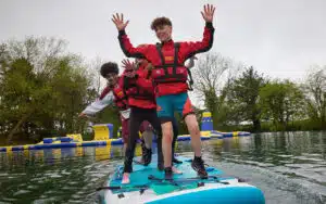 Three people in life vests balance on a paddleboard on a lake, raising their arms. Inflatable structures are visible in the background.