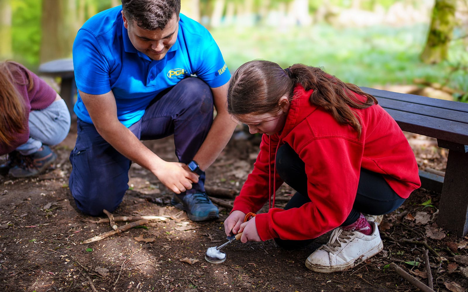 An adult and a child crouch on the ground in a wooded area. The child is using a firestarter tool. A bench and more greenery are visible in the background.