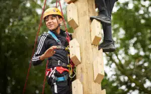Person wearing a helmet and harness climbing a wooden structure outdoors, surrounded by trees.