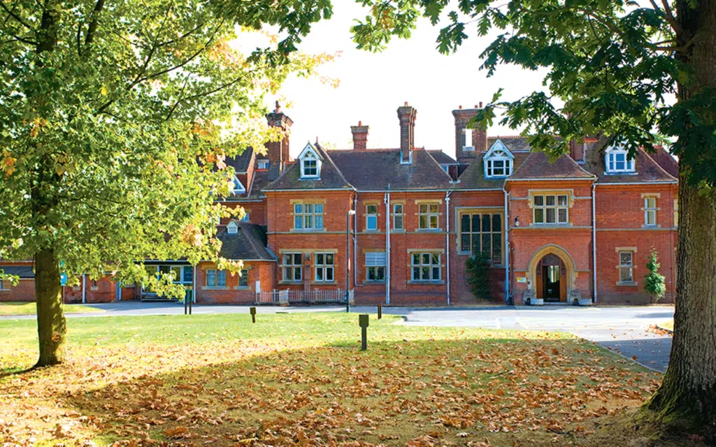 A large red brick building with multiple chimneys and arched entryway, surrounded by trees and a grassy area covered in fallen leaves.