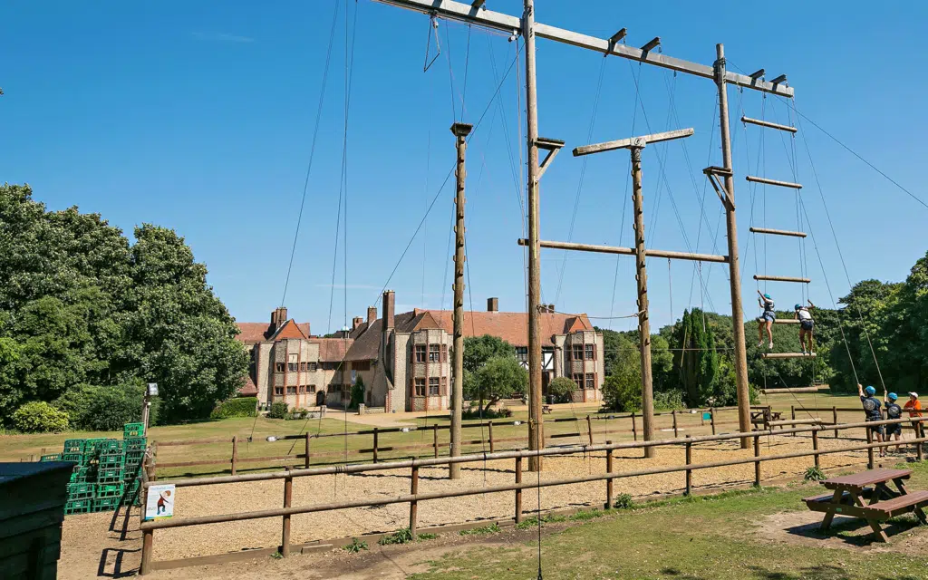 Outdoor ropes course with high poles and climbing ropes. People climbing and standing on platforms. Historic brick building in the background and trees surrounding the area.