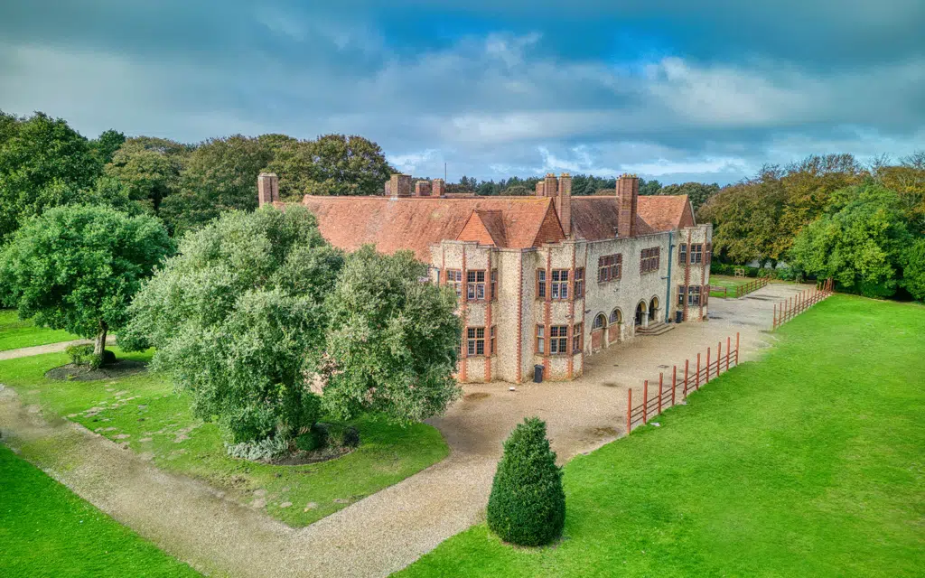 Aerial view of a large, historic brick manor house with multiple chimneys, surrounded by green lawns and trees under a partly cloudy sky.