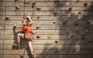 A boy in a red shirt and shorts is engaged in a multi-activity experience, climbing a wooden rock wall with multicolored holds, secured by a harness and rope.