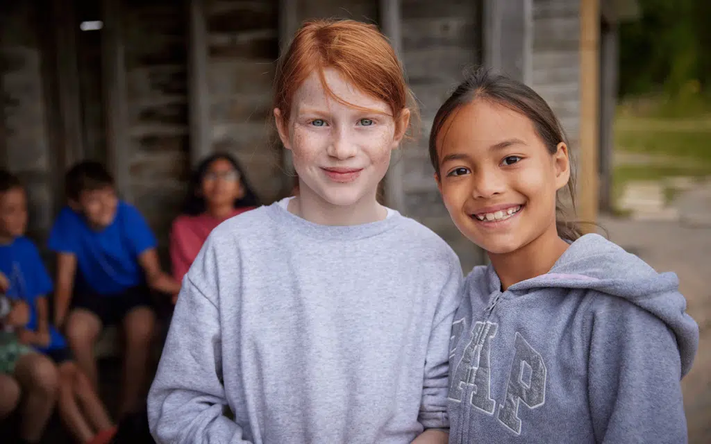Two girls smiling at the camera, standing in front of a wooden structure buzzing with multi-activity. Other children are seated in the background.