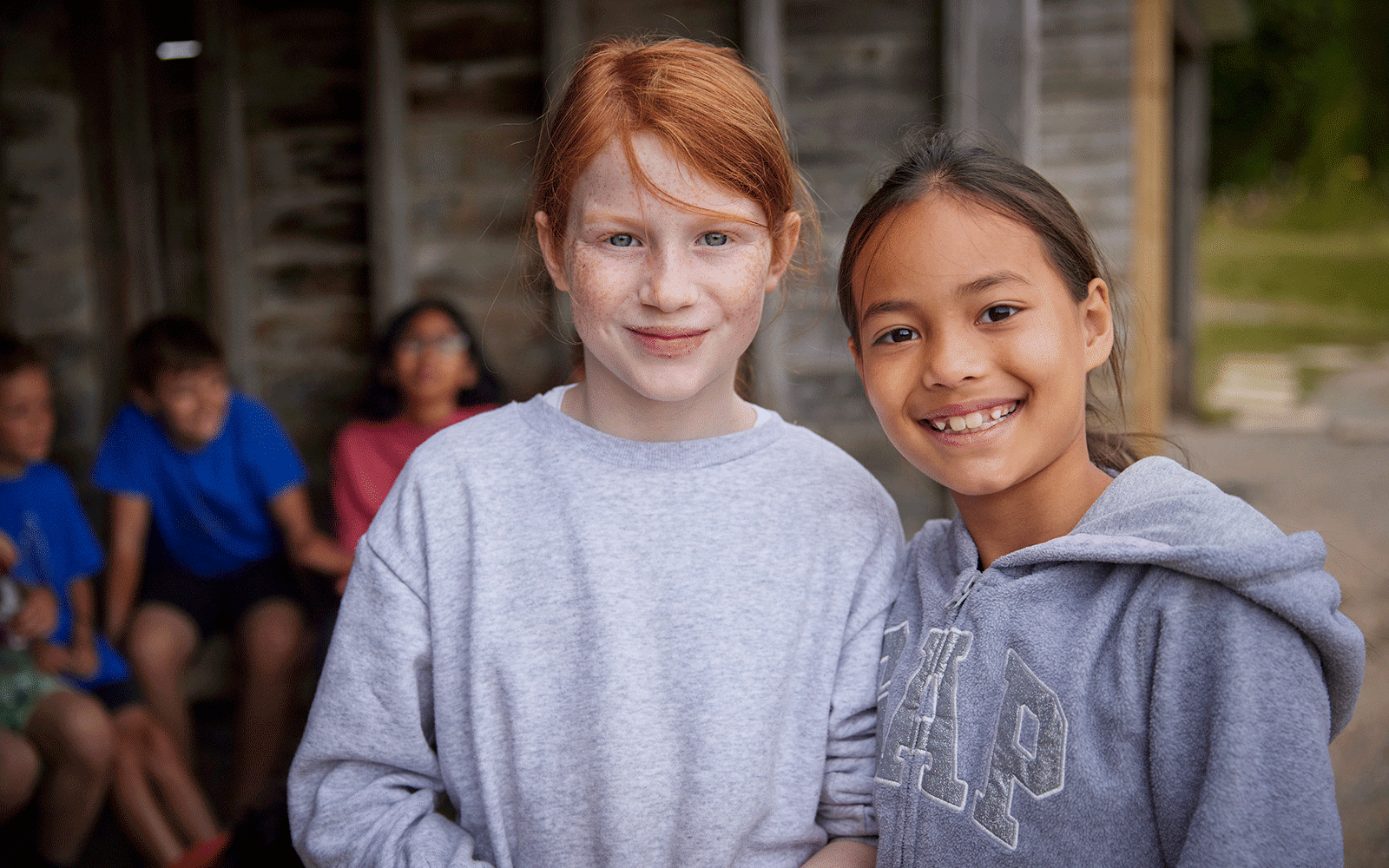 Two girls smiling at the camera, standing in front of a wooden structure buzzing with multi-activity. Other children are seated in the background.