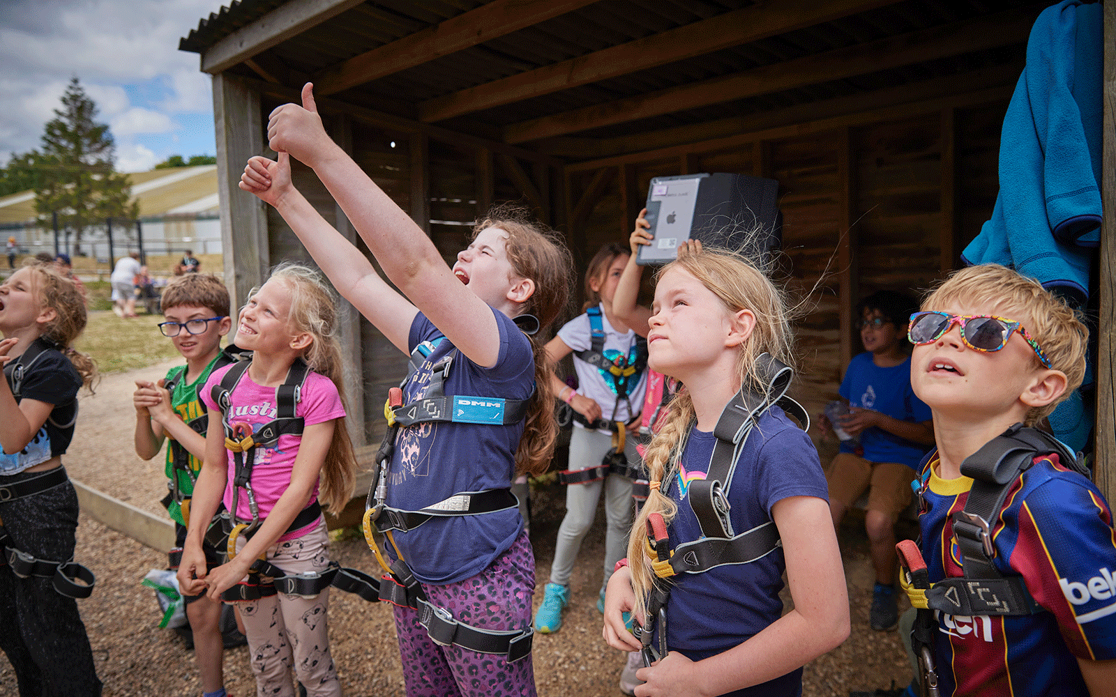 Children in harnesses eagerly watch something overhead, with one girl giving a thumbs-up. They stand outside a wooden shelter on a sunny day, ready for their multi-activity adventure.