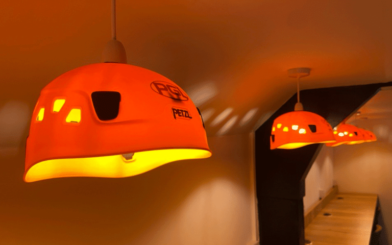 Three orange lampshades made from helmets hang from a ceiling, casting warm light on a wooden shelf below.