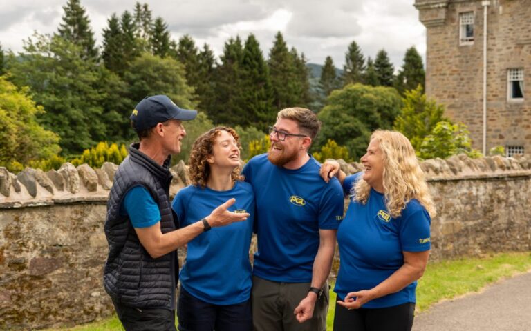 Four smiling people in blue shirts stand outdoors near a stone wall and trees, engaged in conversation.