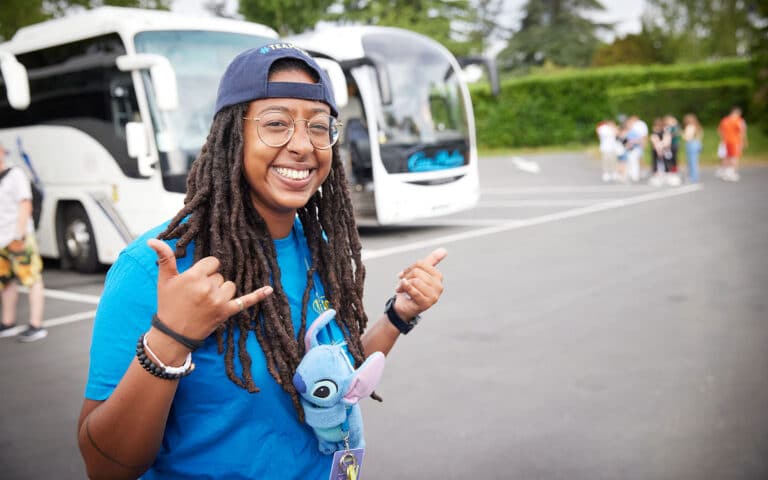 Person with long hair and glasses, wearing a blue shirt and cap, smiles and gestures with hands in front of parked buses. A small plush toy is attached to the shirt.