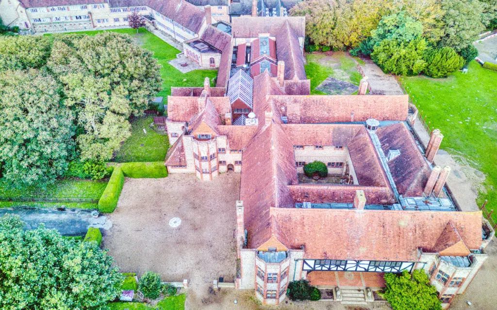Aerial view of a large, historic building with red-tiled roofs and a courtyard, surrounded by trees and grassy areas.