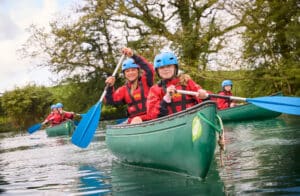 A group of people in red life jackets and blue helmets paddle green canoes on a calm river surrounded by trees.