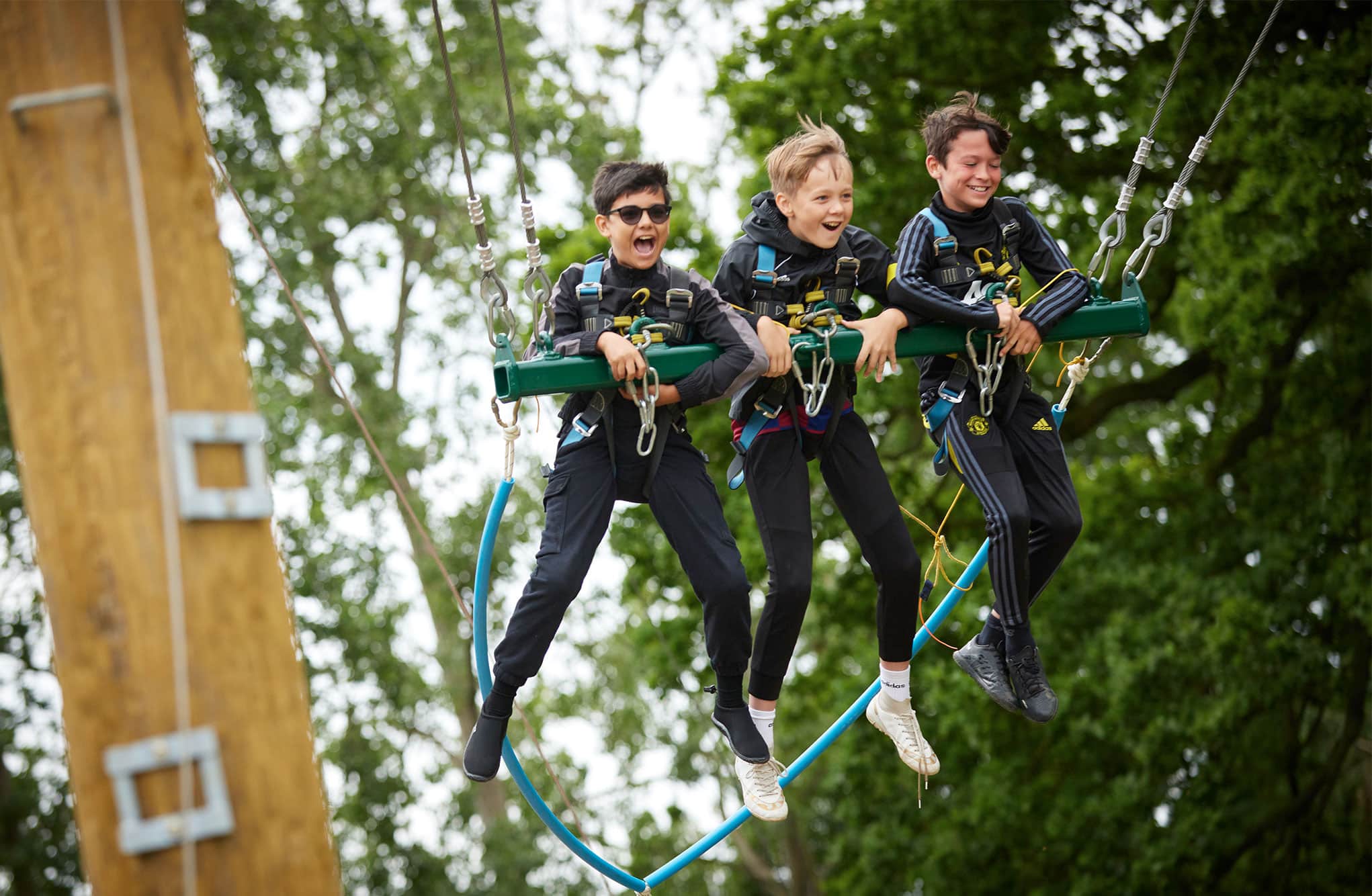 Three boys are harnessed to a giant swing, laughing as they are suspended in the air against a backdrop of trees.