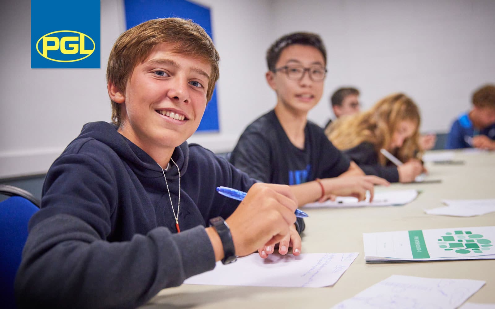 A group of students sit at a table writing in notebooks; one boy in the foreground smiles at the camera. The PGL logo is visible in the top left corner.