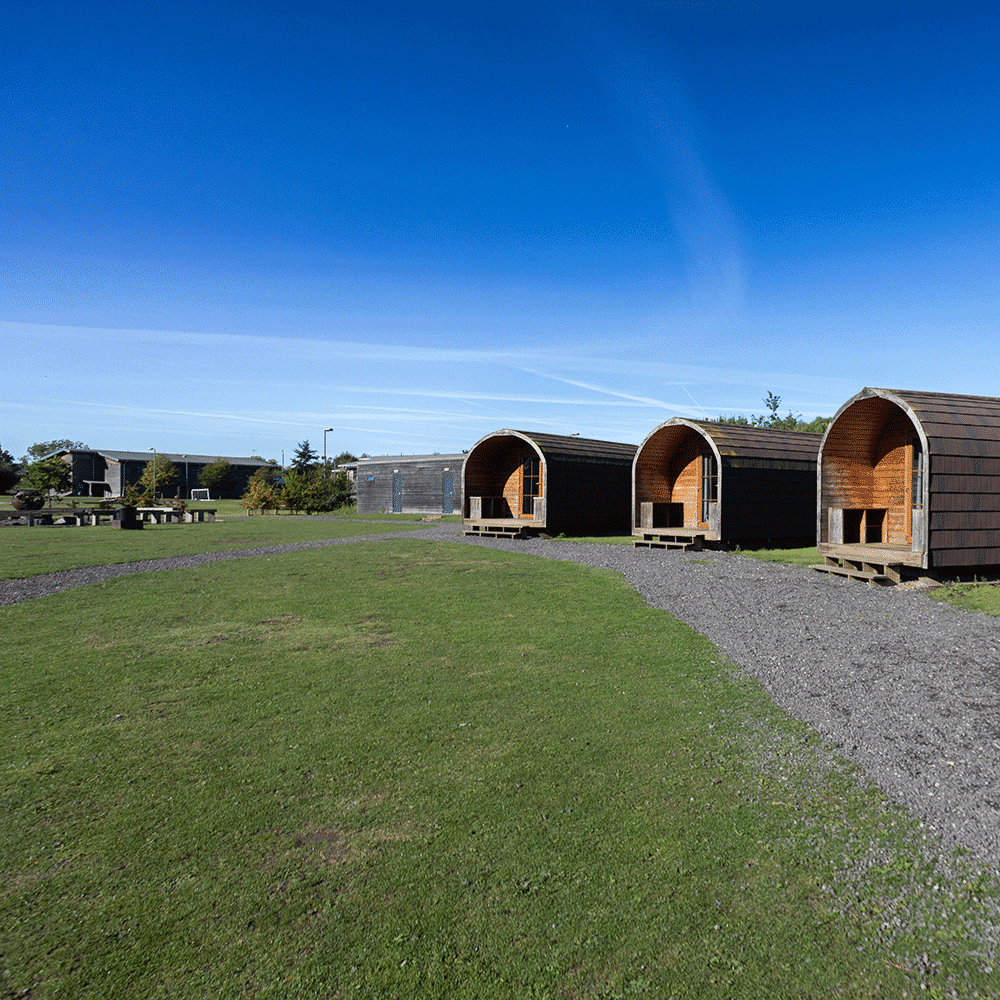 Three wooden camping pods stand in a row on a grassy field under a clear blue sky, with picnic tables and buildings visible in the background.