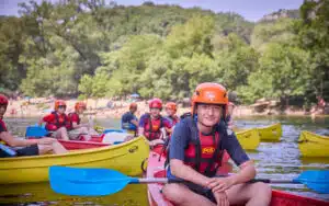 A group of young people wearing helmets and life jackets sit in yellow canoes on a calm river, surrounded by trees, ready for their next multi-activity adventure.