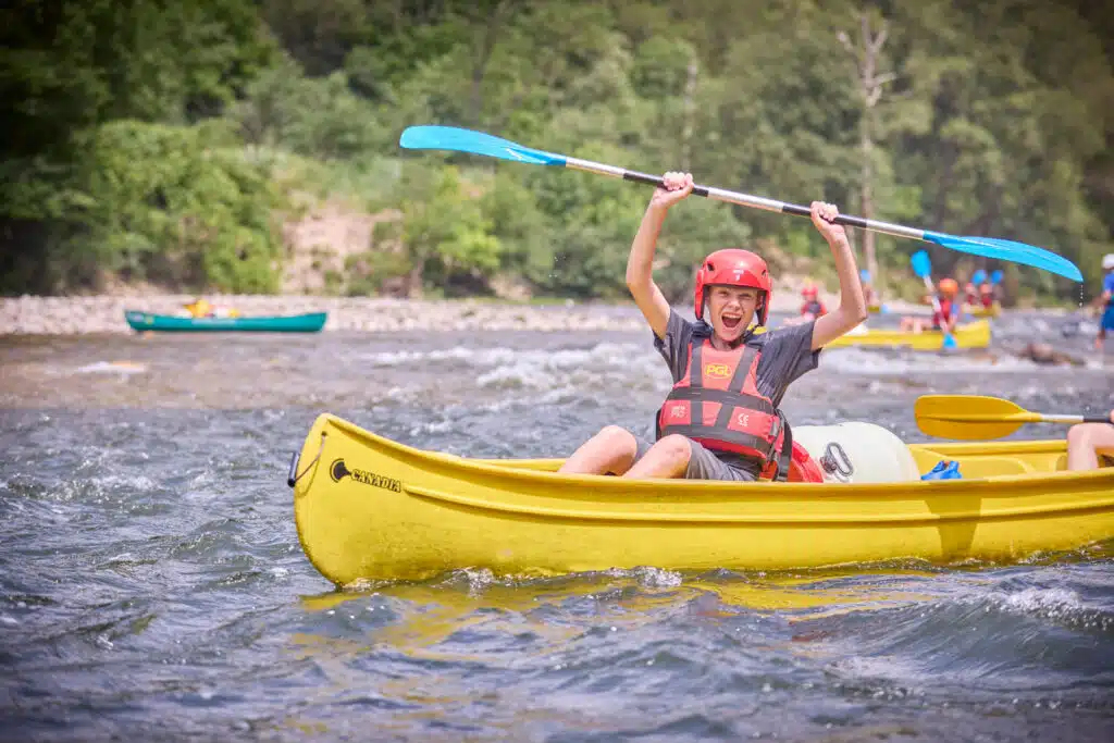 A person wearing a red helmet and life jacket raises a blue paddle while sitting in a yellow kayak on a river with trees in the background, enjoying a multi-activity adventure.