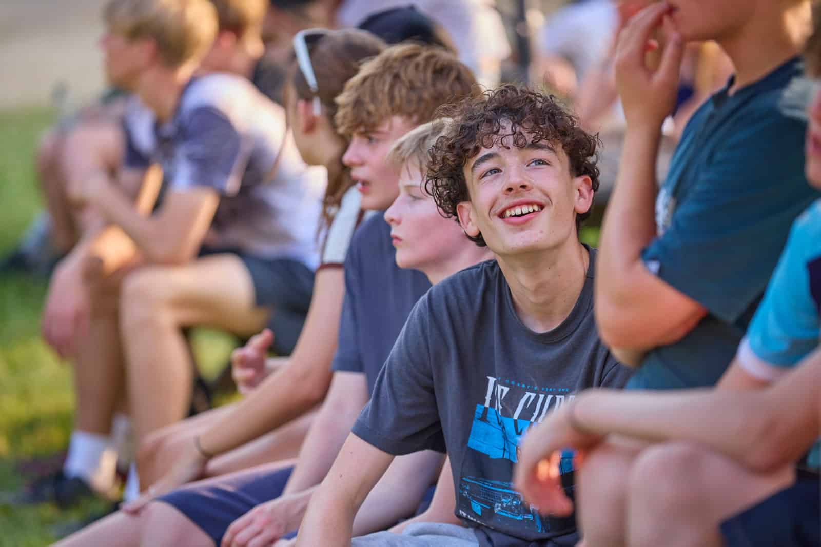 A group of teenagers sit outside on the grass in casual summer clothing, engaged in a multi-activity scene—some chatting while others look ahead, each enjoying the moment.